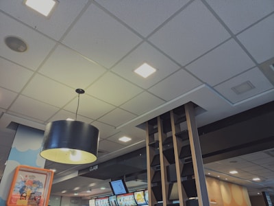 A modern indoor space with a suspended ceiling featuring square recessed lights. A large, black, cylindrical pendant light hangs prominently in the foreground. Digital display screens are visible on the wall, likely showing menus or advertisements. The decor includes contemporary wooden shelving elements and a ceiling-mounted speaker.