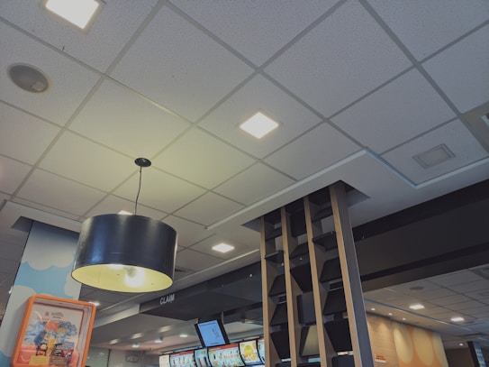 A modern indoor space with a suspended ceiling featuring square recessed lights. A large, black, cylindrical pendant light hangs prominently in the foreground. Digital display screens are visible on the wall, likely showing menus or advertisements. The decor includes contemporary wooden shelving elements and a ceiling-mounted speaker.