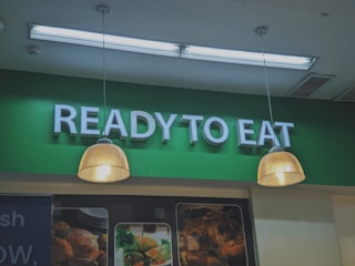 A green sign displaying the words 'READY TO EAT' in large, white letters. Below the sign, there are two hanging lights with cream-toned lampshades. Partially visible food images are displayed beneath the lights.