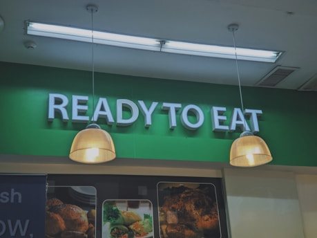 A green sign displaying the words 'READY TO EAT' in large, white letters. Below the sign, there are two hanging lights with cream-toned lampshades. Partially visible food images are displayed beneath the lights.