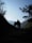 Silhouettes of two hikers walking on a mountain trail surrounded by dense foliage, with a clear sky and distant mountain ranges in the background. The scene is framed by trees and brush on either side, creating a sense of depth and adventure.
