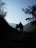 Silhouettes of two hikers walking on a mountain trail surrounded by dense foliage, with a clear sky and distant mountain ranges in the background. The scene is framed by trees and brush on either side, creating a sense of depth and adventure.