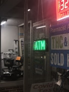 The scene depicts a store window at night with a green ATM sign. Inside, there's a black mobility scooter covered with transparent plastic and marked with a price tag. Surrounding signage includes references to THC, COVID-19 tests, and a lottery jackpot display.