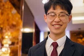 A person wearing glasses and a dark suit with a white shirt and red tie, smiling warmly. The background features a soft, warm light with a golden hue and some abstract shapes, possibly festive decorations.