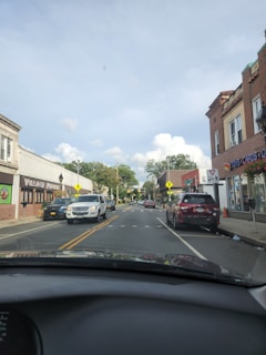 A street view from inside a car shows a small town with parked vehicles on both sides. Buildings have storefronts with signage, including one for toys and cards, and another labeled Village Market. There are street markings, pedestrian crossing signs, and vehicles traveling on the road. The sky is partly cloudy with trees in the background.