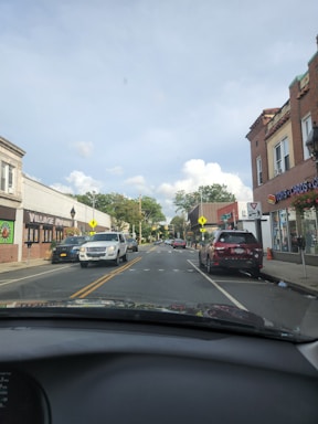 A street view from inside a car shows a small town with parked vehicles on both sides. Buildings have storefronts with signage, including one for toys and cards, and another labeled Village Market. There are street markings, pedestrian crossing signs, and vehicles traveling on the road. The sky is partly cloudy with trees in the background.