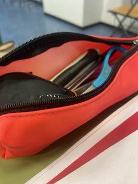 A red pencil case is unzipped, showing various writing instruments inside, including pens and a marker. The background features a blurred setting with a blue floor and pieces of furniture.