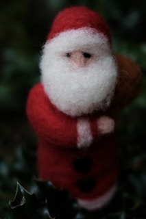 A felted wool figure resembling Santa Claus, complete with a red suit and white beard, standing amidst dark green leaves.