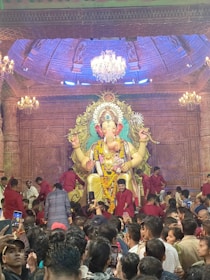 A large, intricately decorated statue of a deity is prominently displayed on a stage, surrounded by an ornate backdrop. Many people, some in red clothing, are gathered around the statue, taking photos and participating in the event. The ceiling is adorned with chandeliers and detailed carvings, contributing to a festive atmosphere.