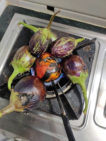 Several round brinjals and one tomato are being roasted directly on a gas stove burner. The vegetables are getting charred and cooked over an open flame, with kitchen utensils visible around the area.