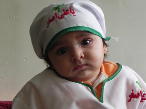 A young child wearing a white and green outfit with red embroidery and a matching white hat. The child has large, expressive eyes and is looking directly at the camera with a neutral expression. The background is a plain wall.