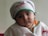 A young child wearing a white and green outfit with red embroidery and a matching white hat. The child has large, expressive eyes and is looking directly at the camera with a neutral expression. The background is a plain wall.