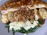 A plate featuring slices of grilled chicken breast topped with a rich brown sauce. Accompanying the chicken are slices of seasoned roasted potatoes and crispy fried onions. Beneath lies a bed of sautéed spinach mixed with melted white cheese, creating a creamy texture.