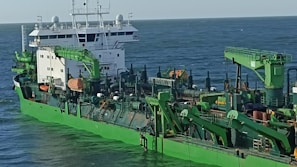 A large industrial ship with green machinery and structures, featuring cranes and various equipment, is shown traveling at sea under a clear sky.