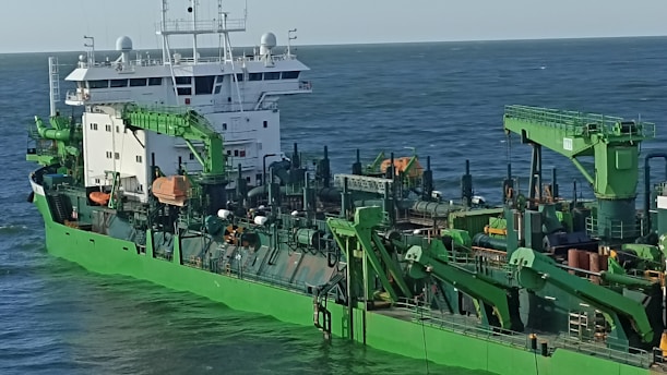 A large industrial ship with green machinery and structures, featuring cranes and various equipment, is shown traveling at sea under a clear sky.