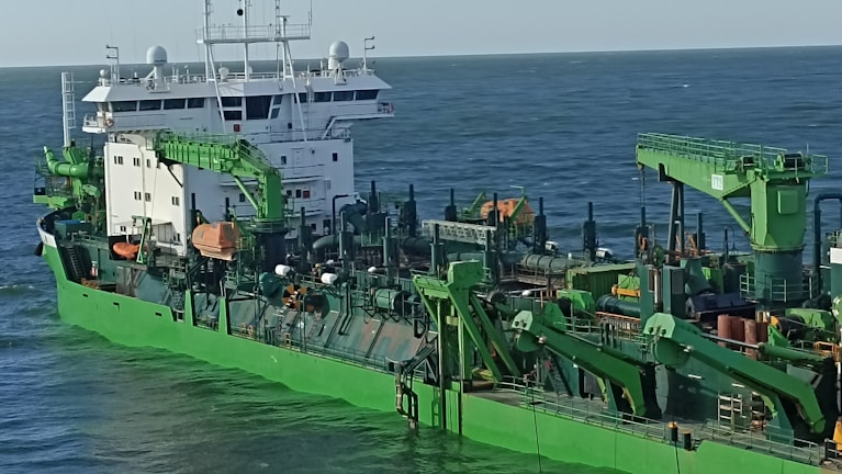 A large industrial ship with green machinery and structures, featuring cranes and various equipment, is shown traveling at sea under a clear sky.