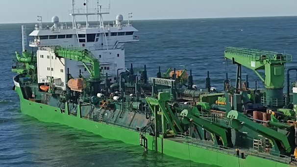 A large industrial ship with green machinery and structures, featuring cranes and various equipment, is shown traveling at sea under a clear sky.