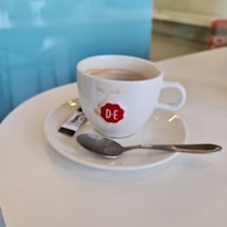 A white ceramic coffee cup with the letters D.E and a red logo is placed on a saucer. Adjacent to the cup is a metal teaspoon, and beside it lies a packet of sugar. The background includes a light blue wall, creating a calm ambiance.