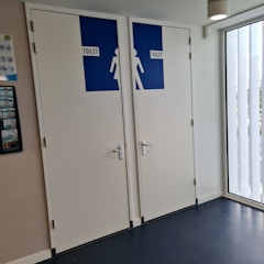 Two restroom doors are located side by side, each with a blue sign indicating the word 'TOILET' and silhouette figures for male and female. The doors are white with silver handles, and the area is well-lit by a ceiling light and natural light from a window. A wall-mounted poster with miscellaneous information is visible on the left.