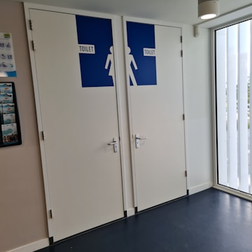 Two restroom doors are located side by side, each with a blue sign indicating the word 'TOILET' and silhouette figures for male and female. The doors are white with silver handles, and the area is well-lit by a ceiling light and natural light from a window. A wall-mounted poster with miscellaneous information is visible on the left.