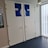 Two restroom doors are located side by side, each with a blue sign indicating the word 'TOILET' and silhouette figures for male and female. The doors are white with silver handles, and the area is well-lit by a ceiling light and natural light from a window. A wall-mounted poster with miscellaneous information is visible on the left.
