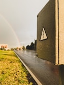 A wet road stretches into the distance, bordered by green grass and a rainbow emerging in the cloudy sky. A roadside utility box, prominently featuring an electrical warning symbol, stands in the foreground. Buildings and trees line the road, and the atmosphere appears calm after rain.