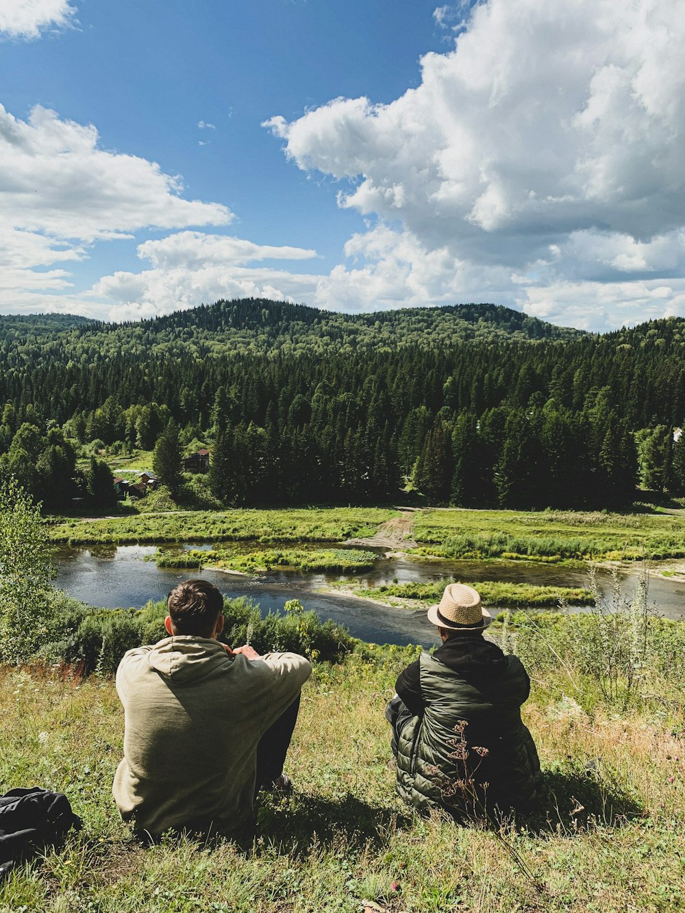 two people sitting on a hill looking at a river and trees