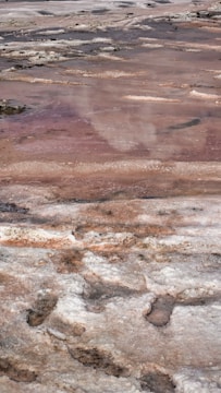 A landscape of salt flats with patches of crystallized, uneven surfaces. Reddish-brown and white tones dominate, with some darker patches scattered throughout, creating a textured and layered appearance.