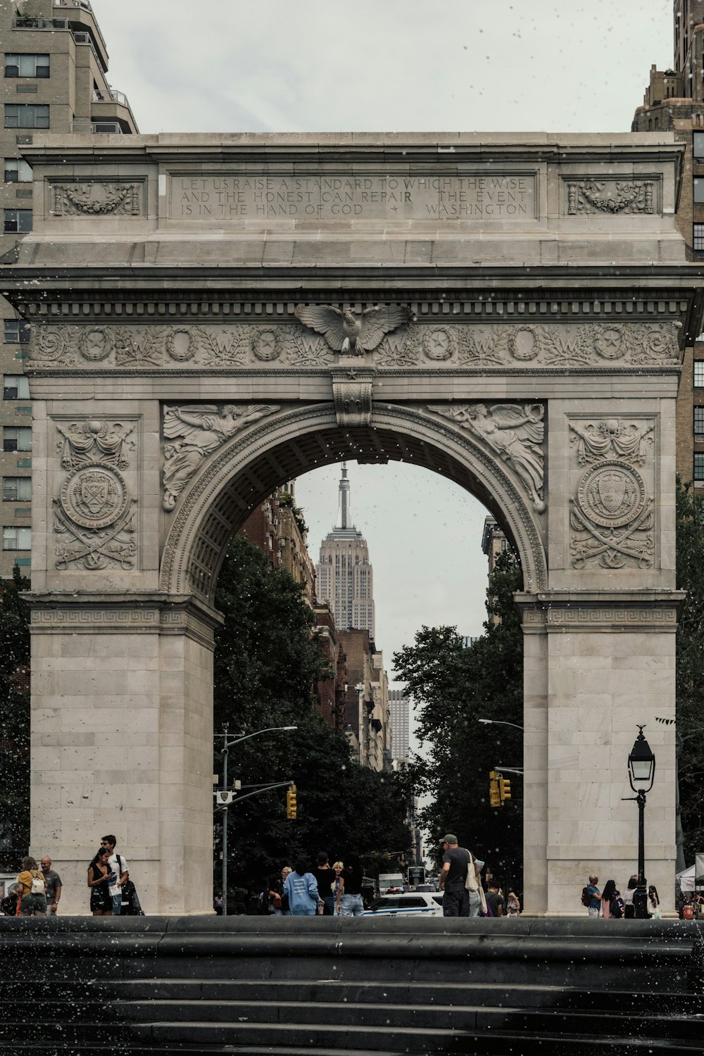 a large stone archway with people walking underneath with Washington Square Park in the background