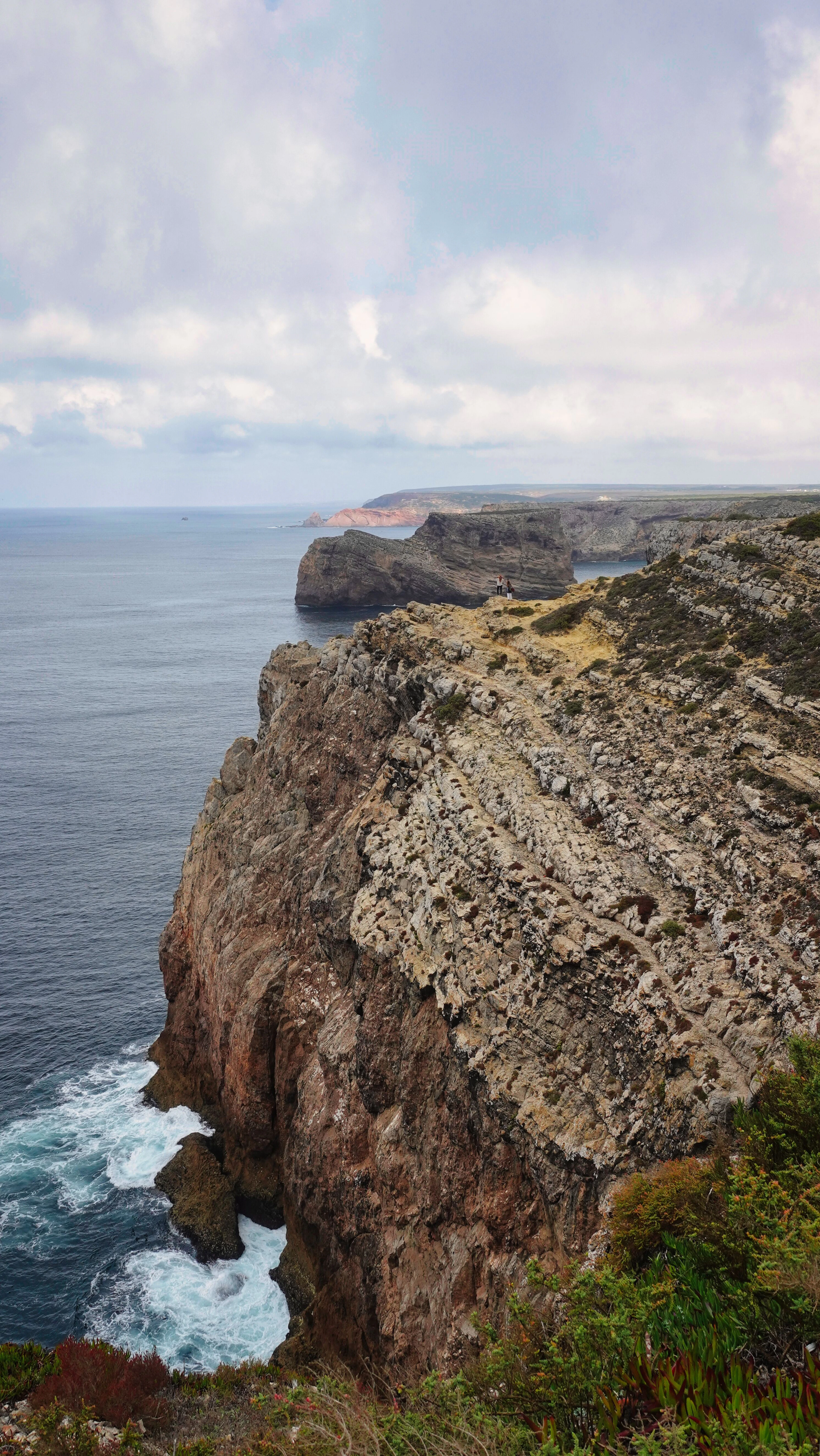 The layered red and white cliffs of Cabo de São Vicente falling sheer into the Atlantic at the southwestern tip of the Algarve, surf breaking white at their foot under heavy cloud.