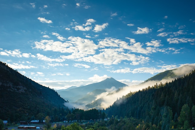 A picturesque mountain landscape with lush green forests and mist rolling through the valleys. The sky is bright and scattered with fluffy white clouds, while a prominent mountain peak stands majestically in the distance.