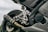 A close-up view of a motorcycle's rear end, showcasing the exhaust, footpeg assembly, and tire. The brand 'Yamaha' is visible on the engine casing. The image emphasizes metal parts and intricate engineering details.
