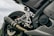 A close-up view of a motorcycle's rear end, showcasing the exhaust, footpeg assembly, and tire. The brand 'Yamaha' is visible on the engine casing. The image emphasizes metal parts and intricate engineering details.