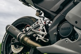 A close-up view of a motorcycle's rear end, showcasing the exhaust, footpeg assembly, and tire. The brand 'Yamaha' is visible on the engine casing. The image emphasizes metal parts and intricate engineering details.