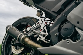 A close-up view of a motorcycle's rear end, showcasing the exhaust, footpeg assembly, and tire. The brand 'Yamaha' is visible on the engine casing. The image emphasizes metal parts and intricate engineering details.