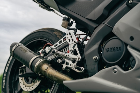 A close-up view of a motorcycle's rear end, showcasing the exhaust, footpeg assembly, and tire. The brand 'Yamaha' is visible on the engine casing. The image emphasizes metal parts and intricate engineering details.