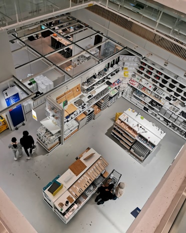 Detailed view of a well-organized store section from an elevated perspective. The image features various household items and cookware arranged on shelves and display tables. Shoppers can be seen browsing through the store. The bright lighting and clean, modern design emphasize the neatness and organization. The store has aisles and designated sections for different types of products.