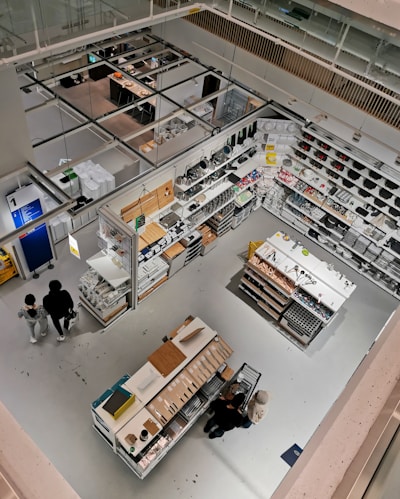 Detailed view of a well-organized store section from an elevated perspective. The image features various household items and cookware arranged on shelves and display tables. Shoppers can be seen browsing through the store. The bright lighting and clean, modern design emphasize the neatness and organization. The store has aisles and designated sections for different types of products.