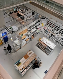 Detailed view of a well-organized store section from an elevated perspective. The image features various household items and cookware arranged on shelves and display tables. Shoppers can be seen browsing through the store. The bright lighting and clean, modern design emphasize the neatness and organization. The store has aisles and designated sections for different types of products.