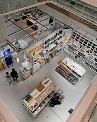 Detailed view of a well-organized store section from an elevated perspective. The image features various household items and cookware arranged on shelves and display tables. Shoppers can be seen browsing through the store. The bright lighting and clean, modern design emphasize the neatness and organization. The store has aisles and designated sections for different types of products.