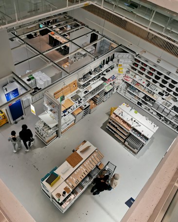 Detailed view of a well-organized store section from an elevated perspective. The image features various household items and cookware arranged on shelves and display tables. Shoppers can be seen browsing through the store. The bright lighting and clean, modern design emphasize the neatness and organization. The store has aisles and designated sections for different types of products.