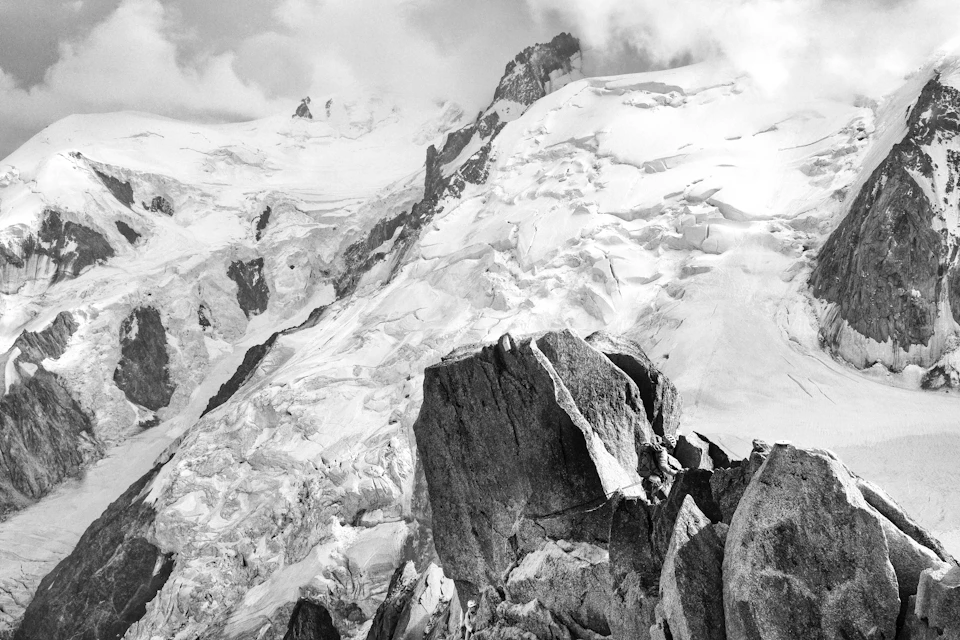 The image depicts a majestic mountain range with snow-capped peaks and a rocky cliff face, all rendered in black and white. The mountains are covered in a thick layer of snow, creating a stark contrast against their dark forms. The perspective is from the top of one of the mountains, looking down towards the base of another mountain. The image conveys a sense of grandeur and scale, emphasizing the vastness and beauty of nature's creations.