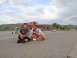 Two people are sitting on a paved area with a large sign in the background that reads 'Kuta Mandalika.' The sky is partly cloudy, and there is a mountainous landscape in the distance. There are some trees, a green trash bin, and other people walking in the background.