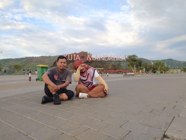 Two people are sitting on a paved area with a large sign in the background that reads 'Kuta Mandalika.' The sky is partly cloudy, and there is a mountainous landscape in the distance. There are some trees, a green trash bin, and other people walking in the background.