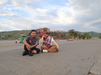 Two people are sitting on a paved area with a large sign in the background that reads 'Kuta Mandalika.' The sky is partly cloudy, and there is a mountainous landscape in the distance. There are some trees, a green trash bin, and other people walking in the background.