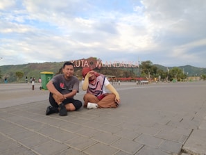 Two people are sitting on a paved area with a large sign in the background that reads 'Kuta Mandalika.' The sky is partly cloudy, and there is a mountainous landscape in the distance. There are some trees, a green trash bin, and other people walking in the background.