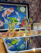 A tabletop scene with a framed artwork depicting colorful fish against a blue background surrounded by floral patterns. In front, there's a glass jar containing a green plant, several containers of paint, and a glass holding an assortment of pens and brushes. A small clock displaying the time sits to the right. The wood surface reflects part of the scene, including the plant and art.