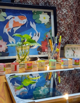 A tabletop scene with a framed artwork depicting colorful fish against a blue background surrounded by floral patterns. In front, there's a glass jar containing a green plant, several containers of paint, and a glass holding an assortment of pens and brushes. A small clock displaying the time sits to the right. The wood surface reflects part of the scene, including the plant and art.