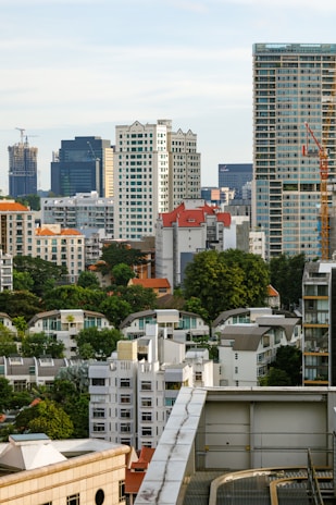 A cityscape featuring a mix of tall modern skyscrapers and smaller residential buildings, some with red tile roofs. Trees and greenery are interspersed among the buildings, and a construction crane is visible, indicating ongoing development. The sky is a light blue with some clouds.