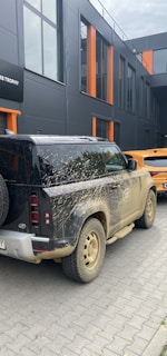 A muddy black off-road vehicle is parked on a paved road next to a modern building. The building features large windows with orange accents.